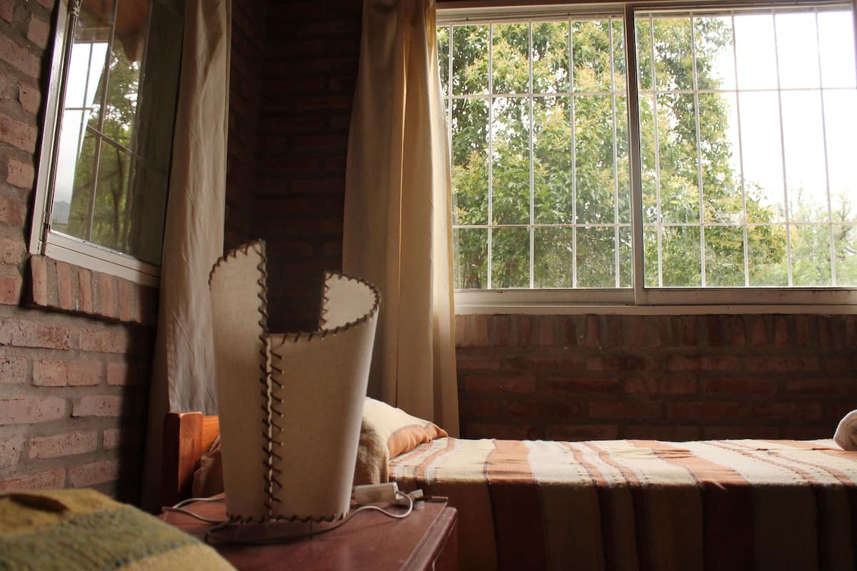 A room with a brick wall, a window with a view of trees, a bed with a striped blanket, and a lamp on a wooden table.