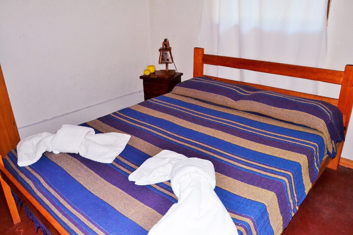 A bed with a striped bedspread, a wooden headboard, and a nightstand with a lamp and a fruit bowl.