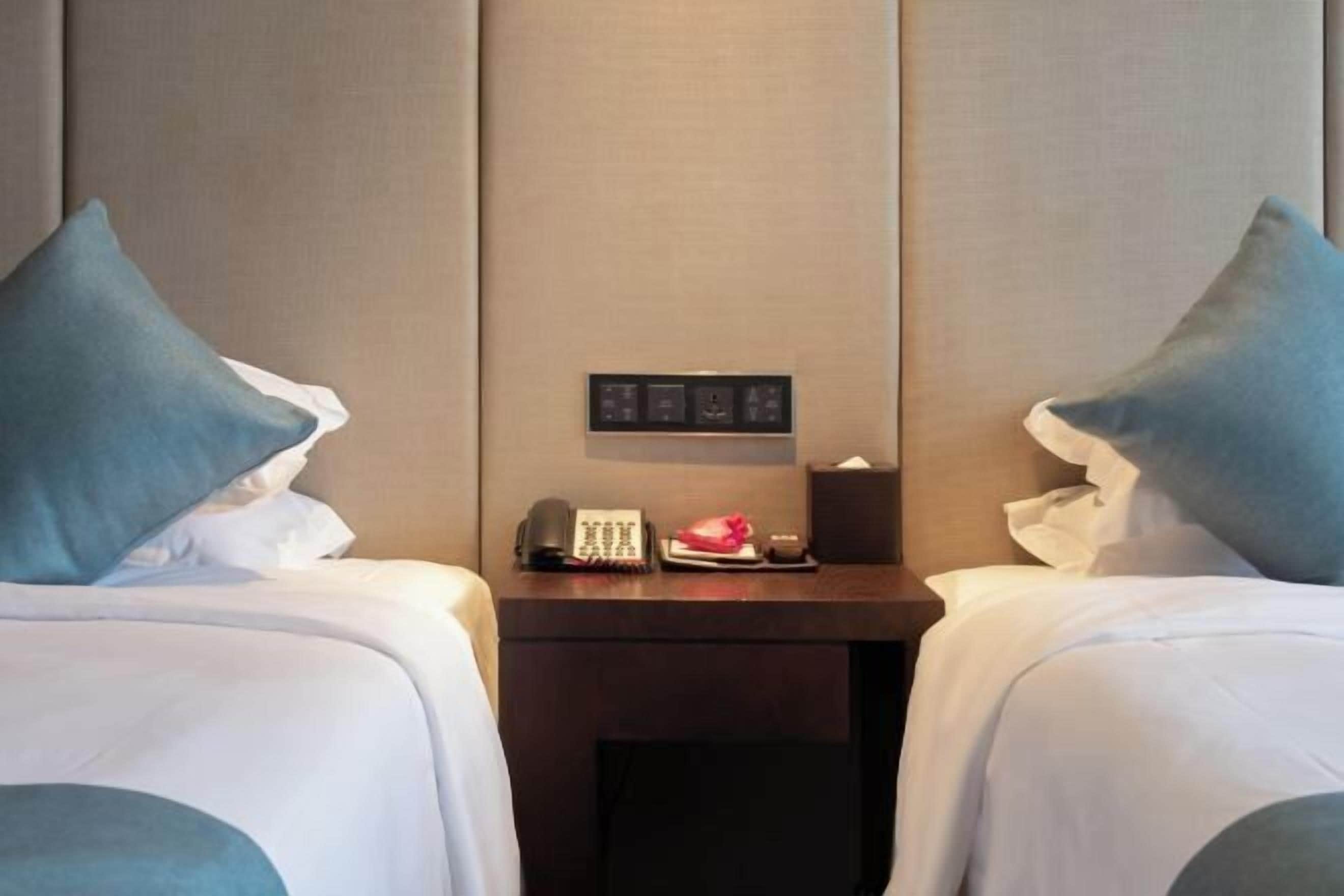 Ein Hotelzimmer mit zwei Betten, einem Nachttisch mit Telefon und Lampe sowie einer wandmontierten Bedienkonsole.