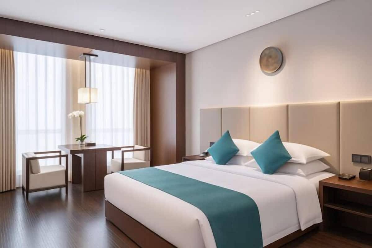 Habitación de hotel con cama, mesita de noche, lámpara, sillón y vistas al exterior a través de amplias ventanas.