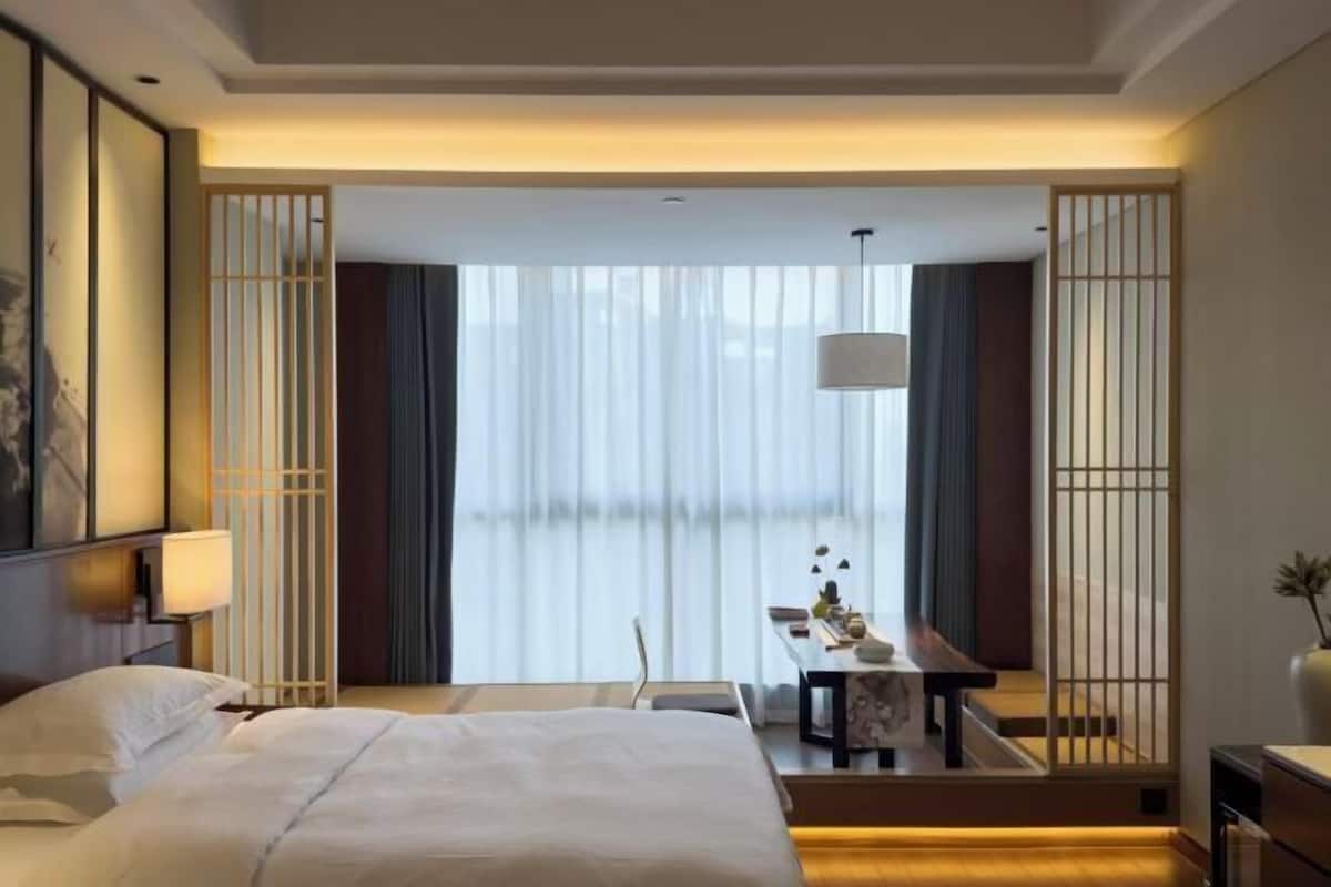 A hotel room with a bed, bedside table, lamp, and a view through a window with curtains.