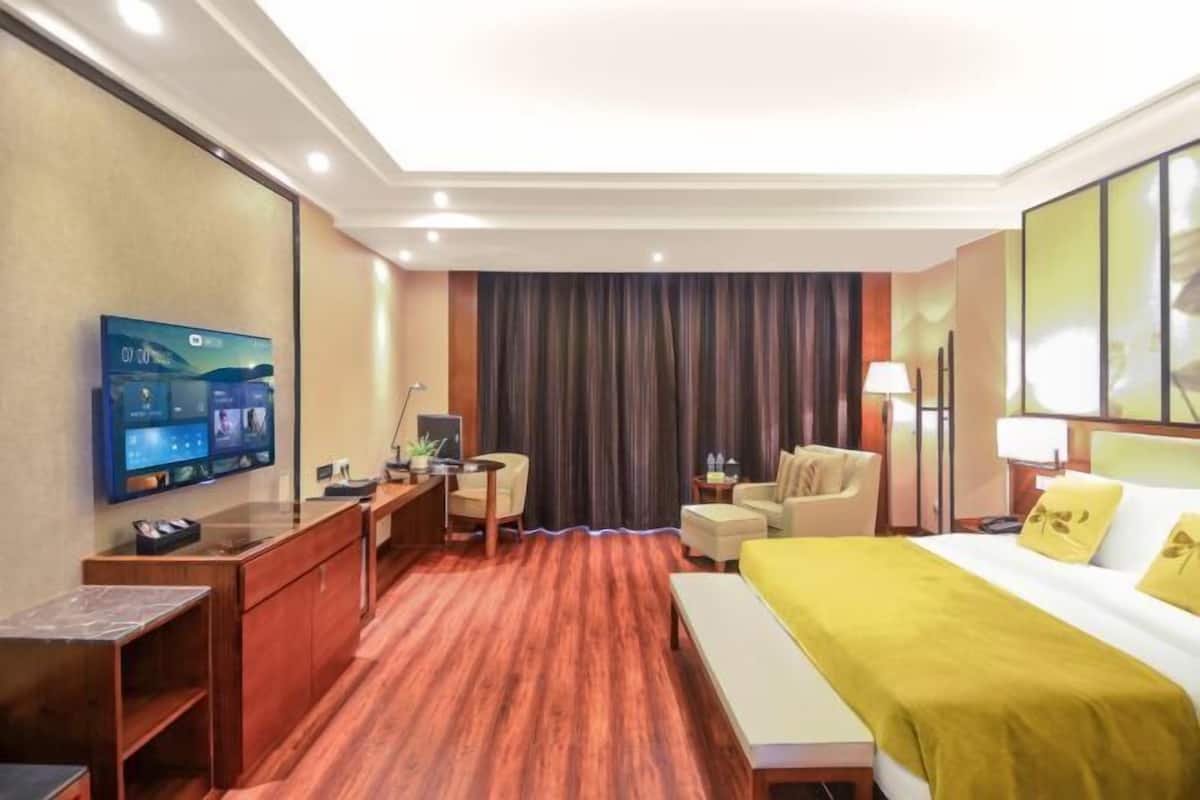 A hotel room with a wooden floor, a bed with a yellow bedspread, a desk with a computer, a chair, a TV mounted on the wall, and a large window with curtains.