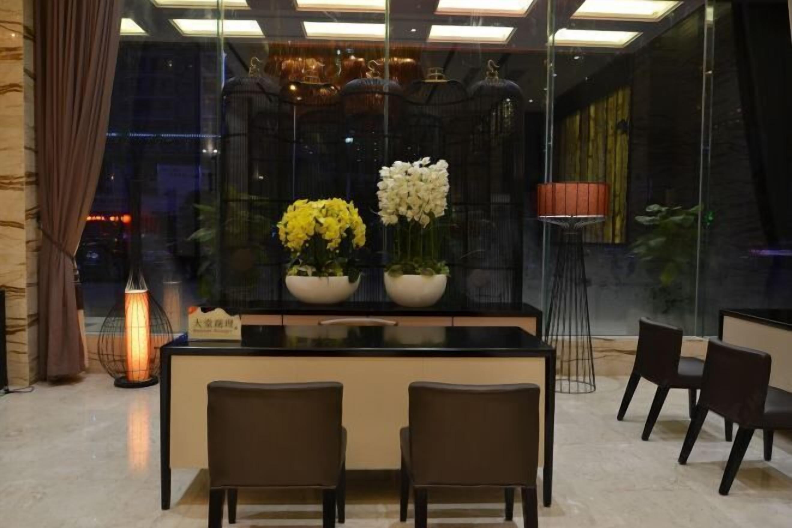 A modern hotel lobby with a reception desk, two chairs, and a large window with decorative plants.