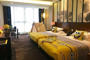 A hotel room with two beds, each covered with yellow SpongeBob-themed bedding. There is a desk with a chair, a lamp, and a large window with curtains. - Xiangtan Mingyi Hotel (Bantang City Railway Station Store) (Xiangtan)