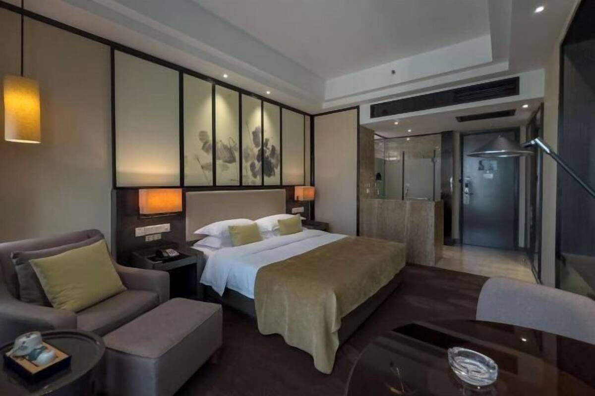 A modern hotel room with a bed, a sofa, a desk, and a glass door leading to a bathroom.