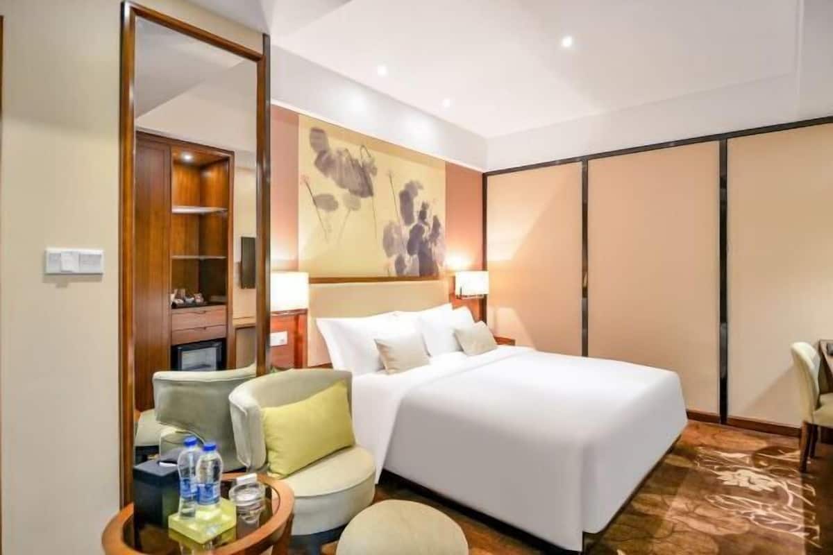 A hotel room with a bed, a chair, a table with water bottles, a mirror, and a wall with a floral painting.