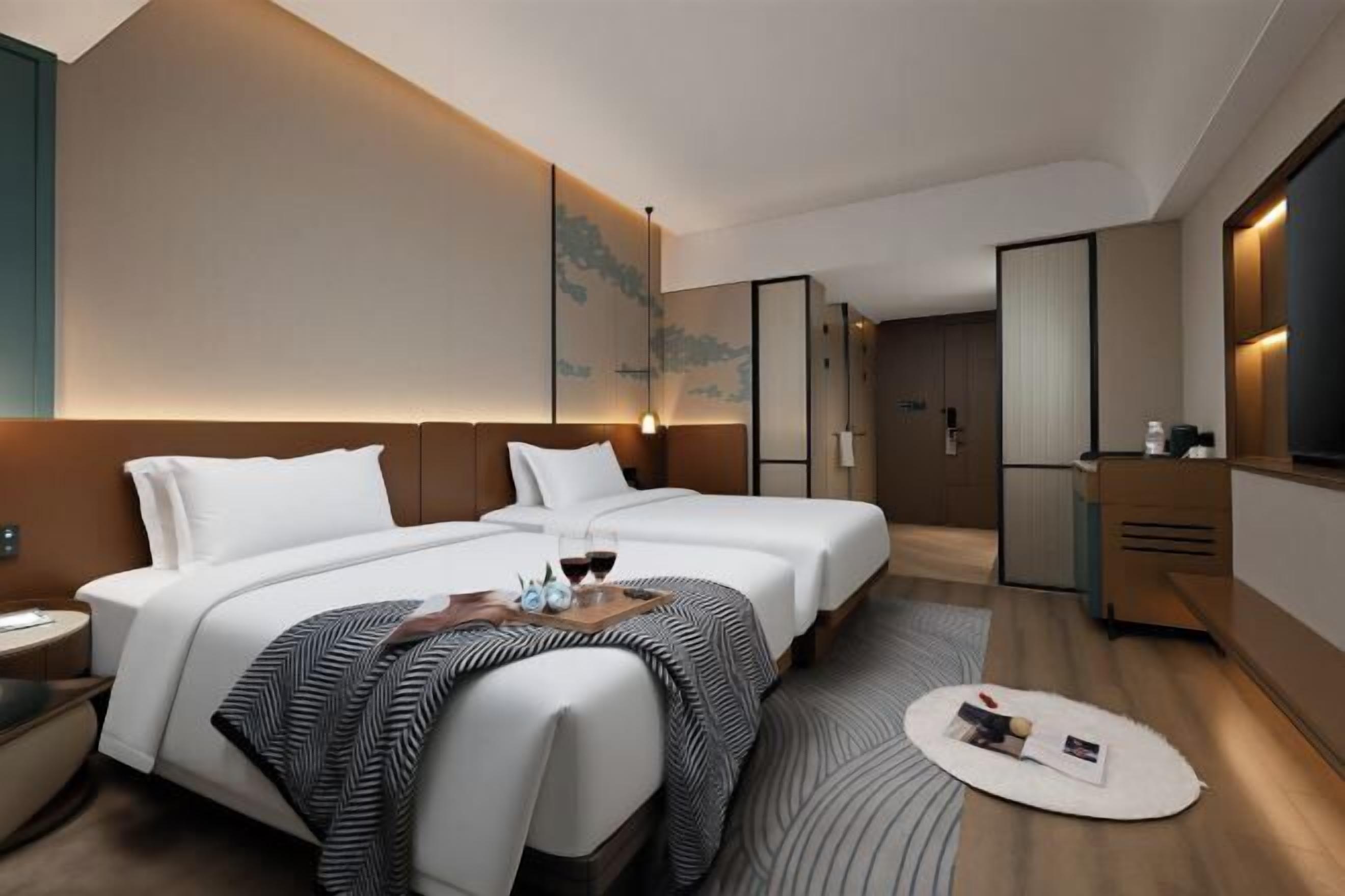 A modern hotel room with two beds, a desk, a mirror, and a view of the city.