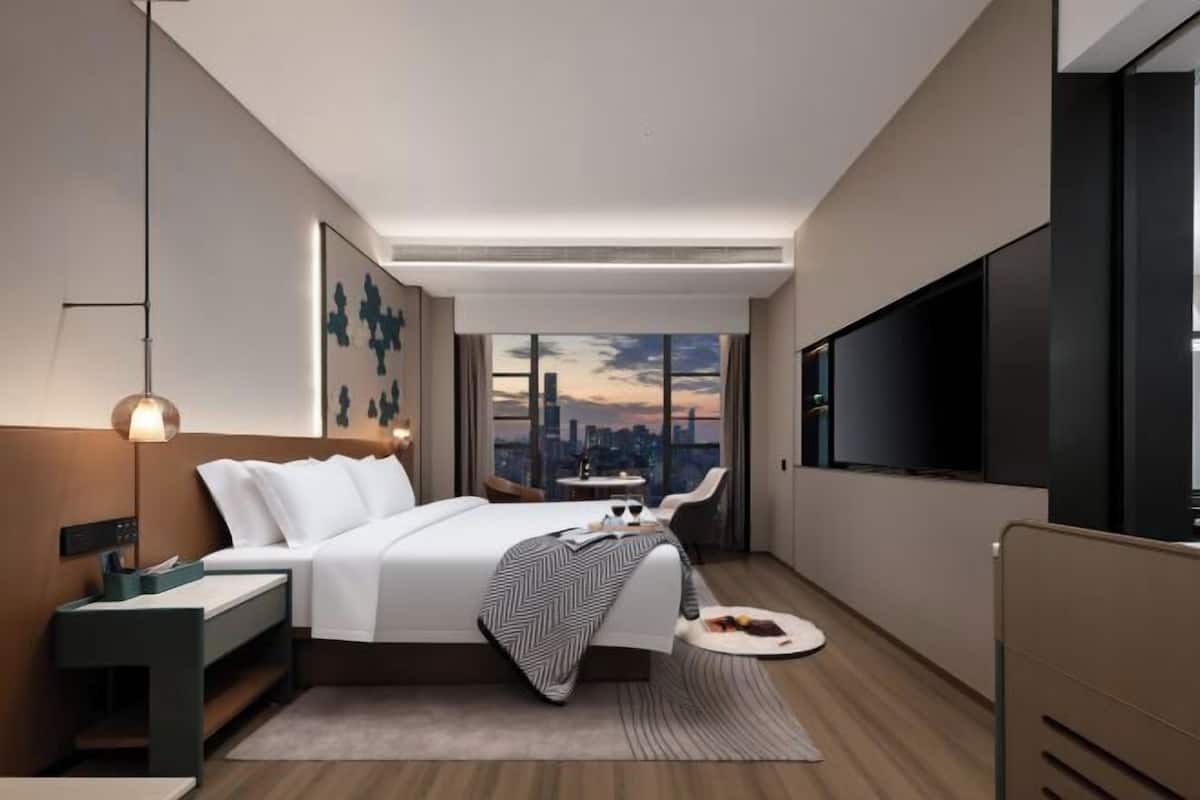 A modern hotel room with a bed, bedside table, lamp, TV, and a city view.