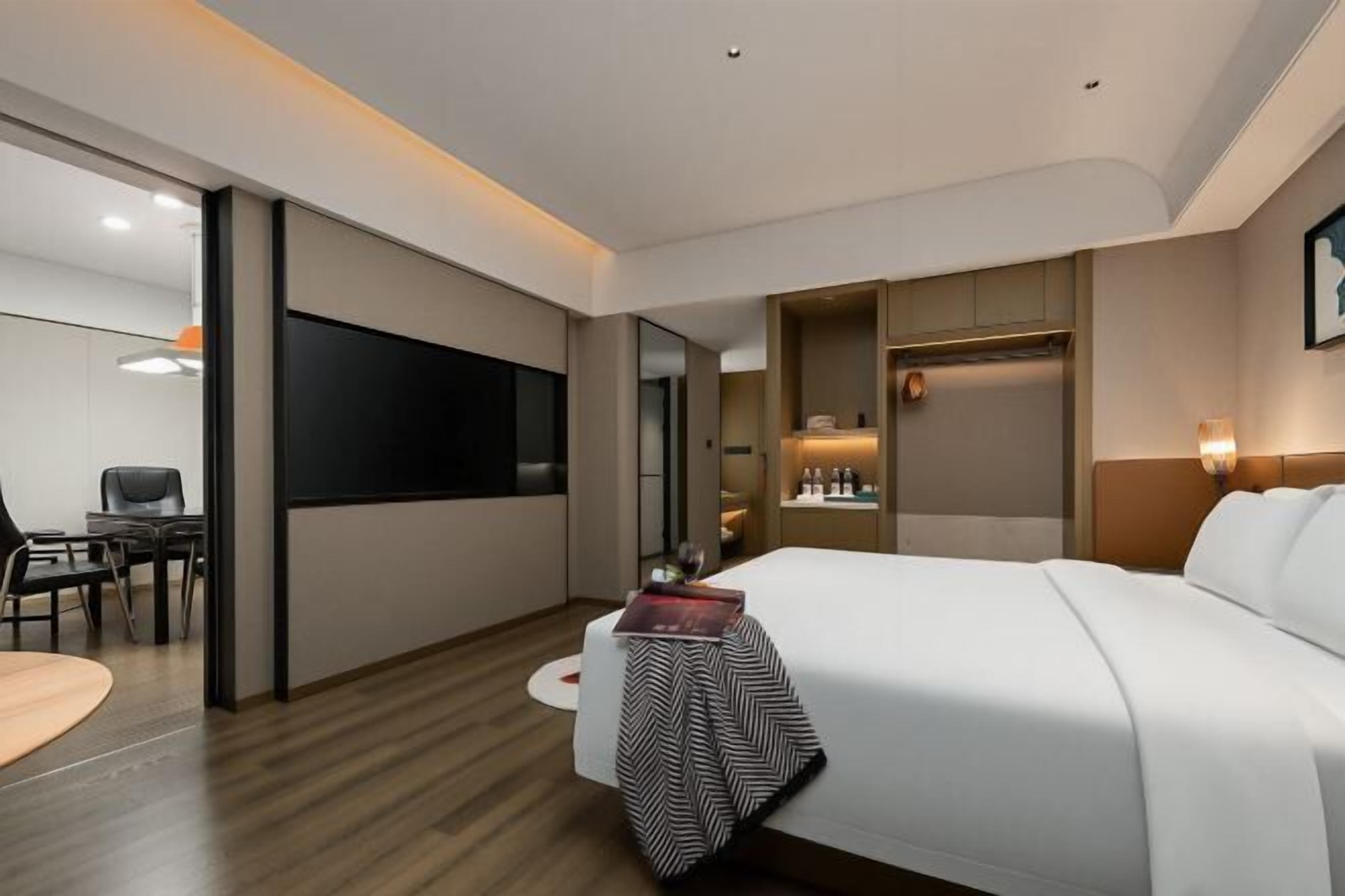 A modern hotel room with a large bed, a desk, a flat-screen TV, and a minibar.