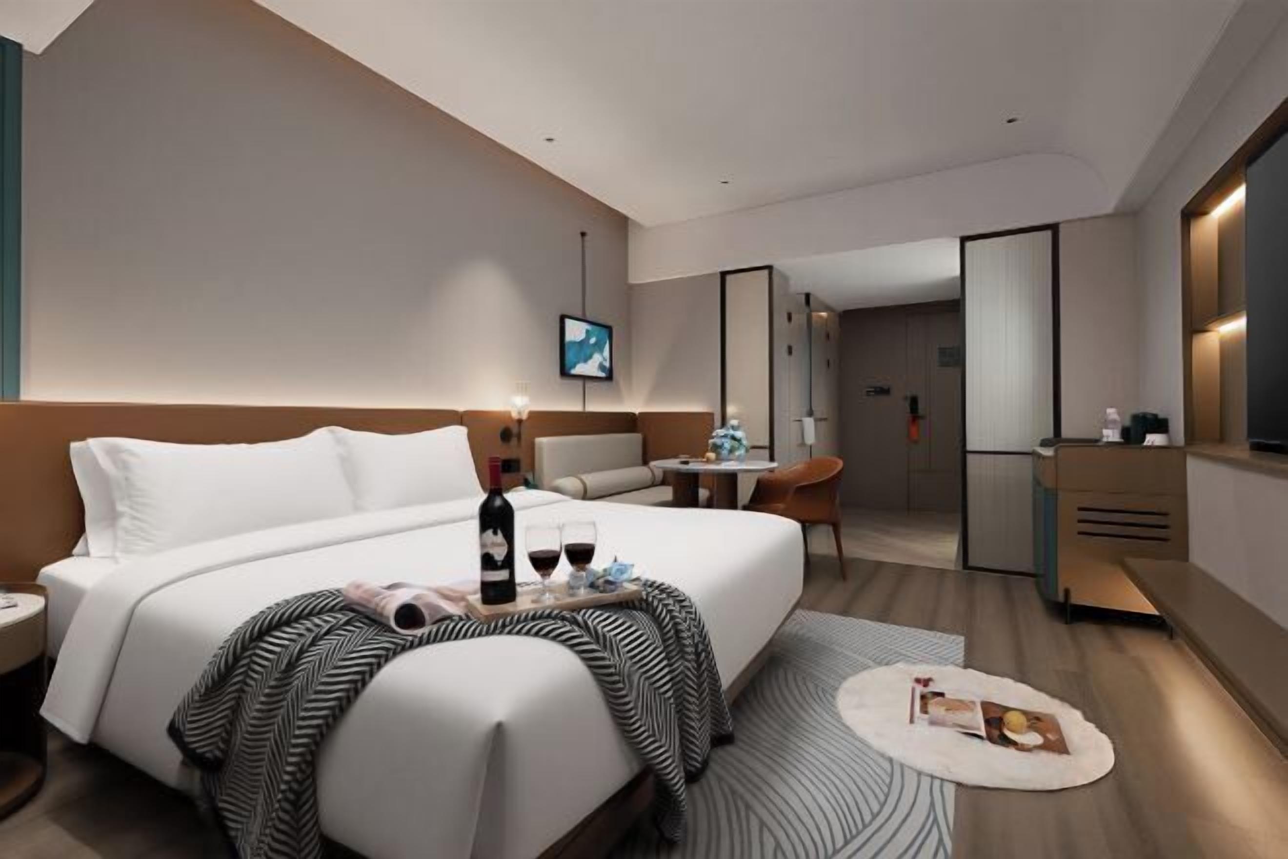 A modern hotel room with a large bed, bedside table, a small desk, a chair, a TV, and a mirror.