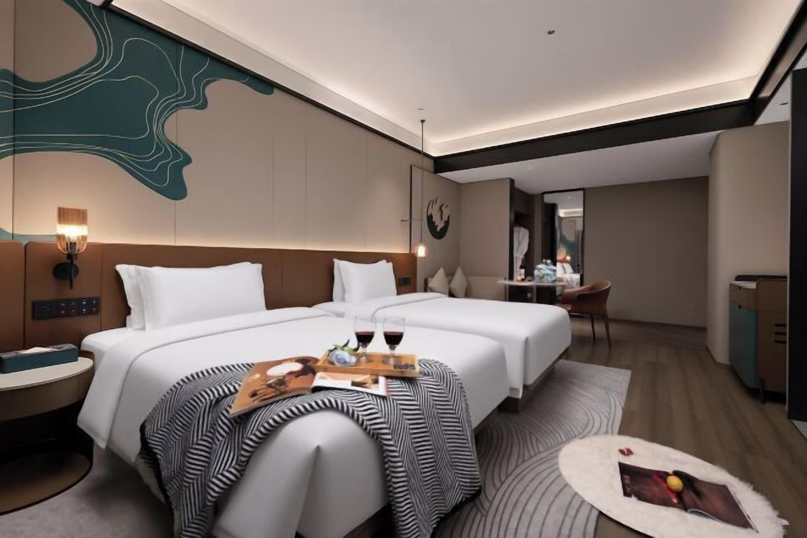 A hotel room with two beds, a desk, a chair, a small table with a tray of wine glasses, and a decorative wall piece.