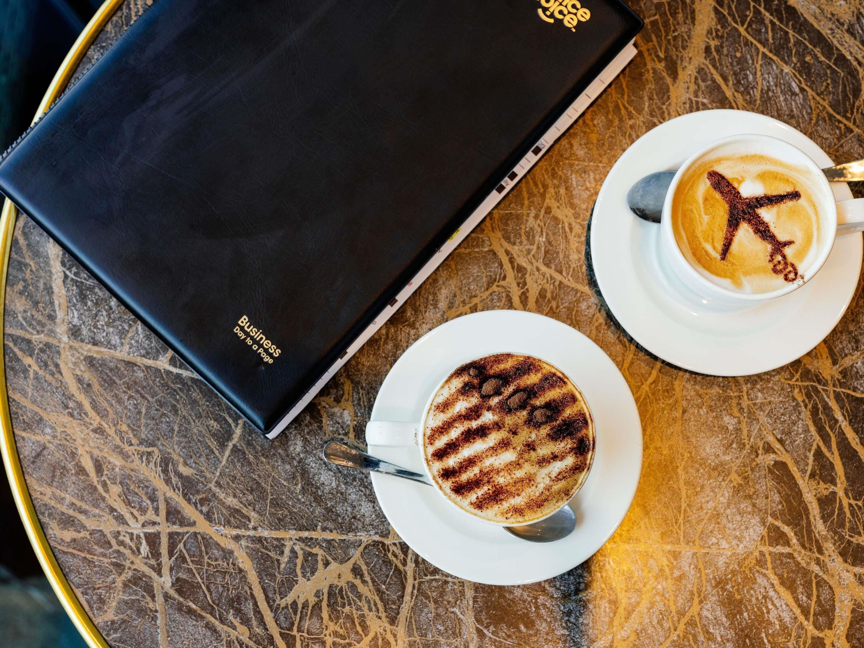 Kannettava tietokone, jonka kannessa on liikemaailmaan liittyvä kuvio, kuppi kahvia, jossa on latte-kuvio, ja aluslautanen marmoripöydällä.