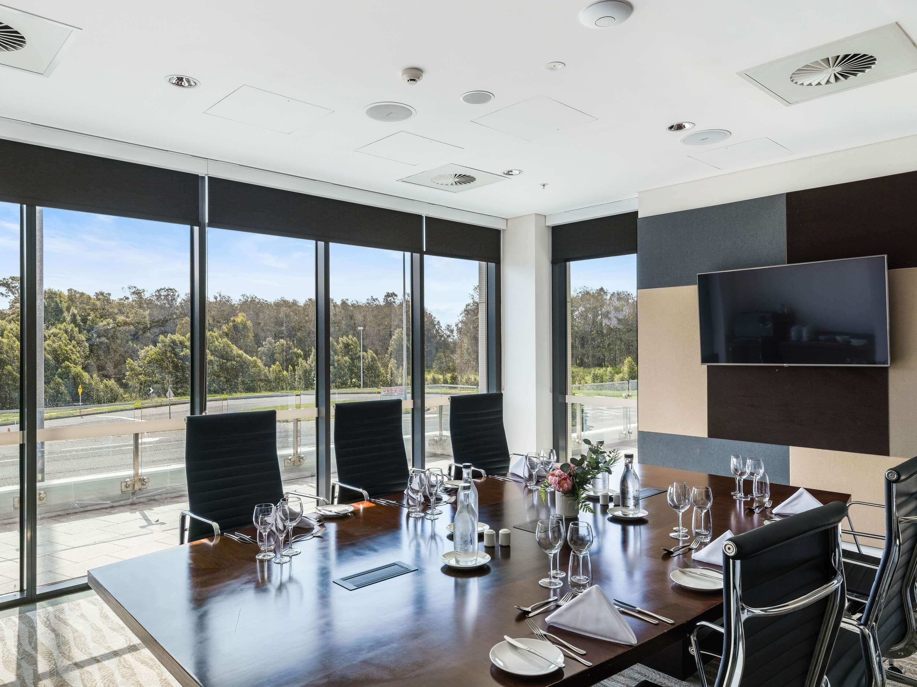 Un salón de conferencias con una gran mesa dispuesta para una reunión, ventanas amplias con vistas a árboles y una carretera, y un televisor montado en la pared.