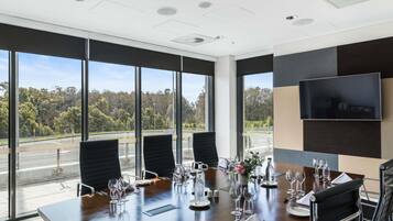 A conference room with a large table set for a meeting, large windows with a view of trees and a road, and a mounted television on the wall.