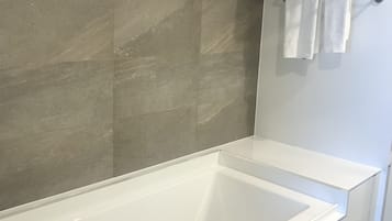 A modern bathroom with a large white bathtub, grey tiled walls, and a towel rack with folded towels.