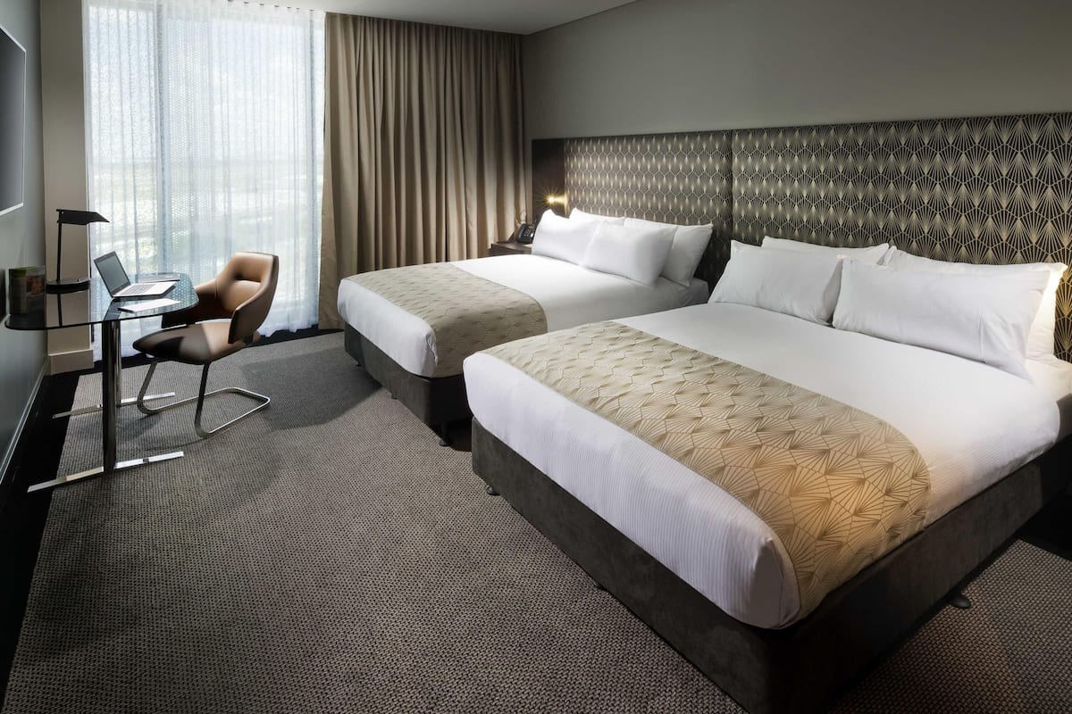 Hotellihuone, jossa on kaksi sänkyä, työpöytä kannettavalla tietokoneella, tuoli ja suuri ikkuna, jossa on verhot.