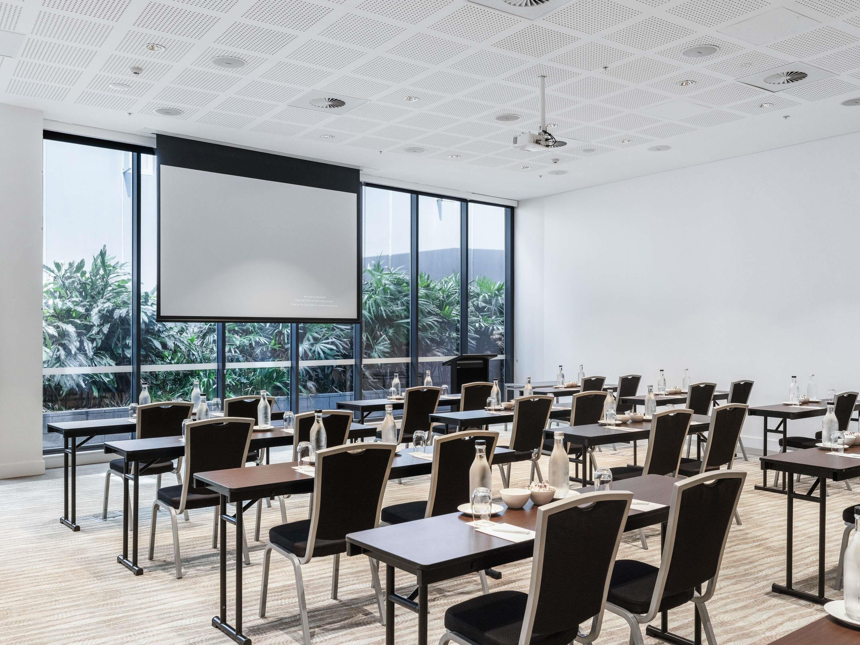 A conference room with a large screen, tables, chairs, and a view of greenery outside.