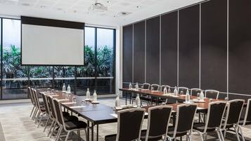 A conference room with a large screen, multiple tables, chairs, and a view of greenery through large windows.