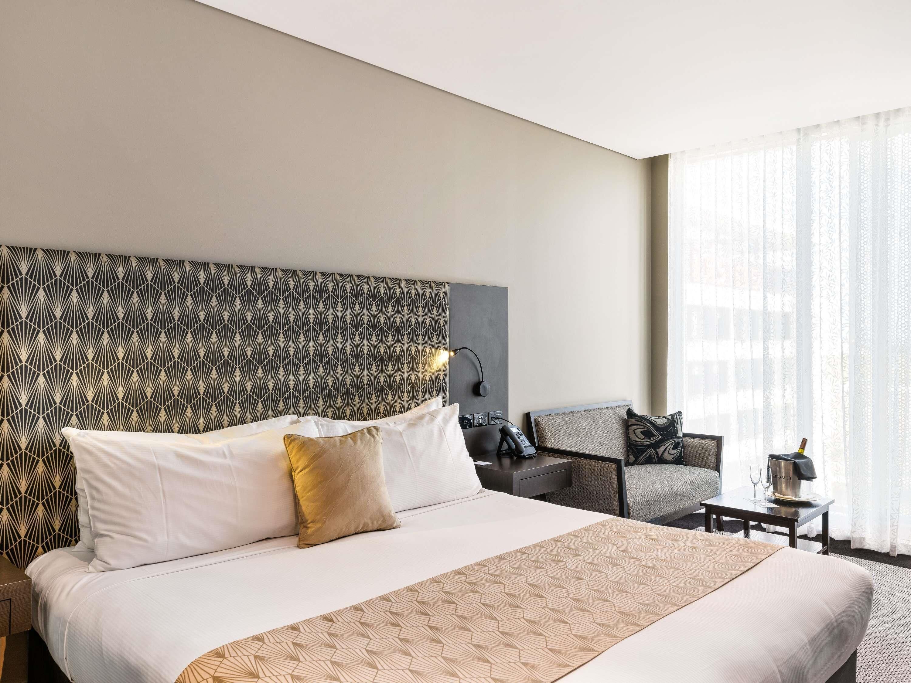 A hotel room with a large bed, a headboard with a geometric pattern, a chair, a small table with a bottle and glasses, and a window with sheer curtains.