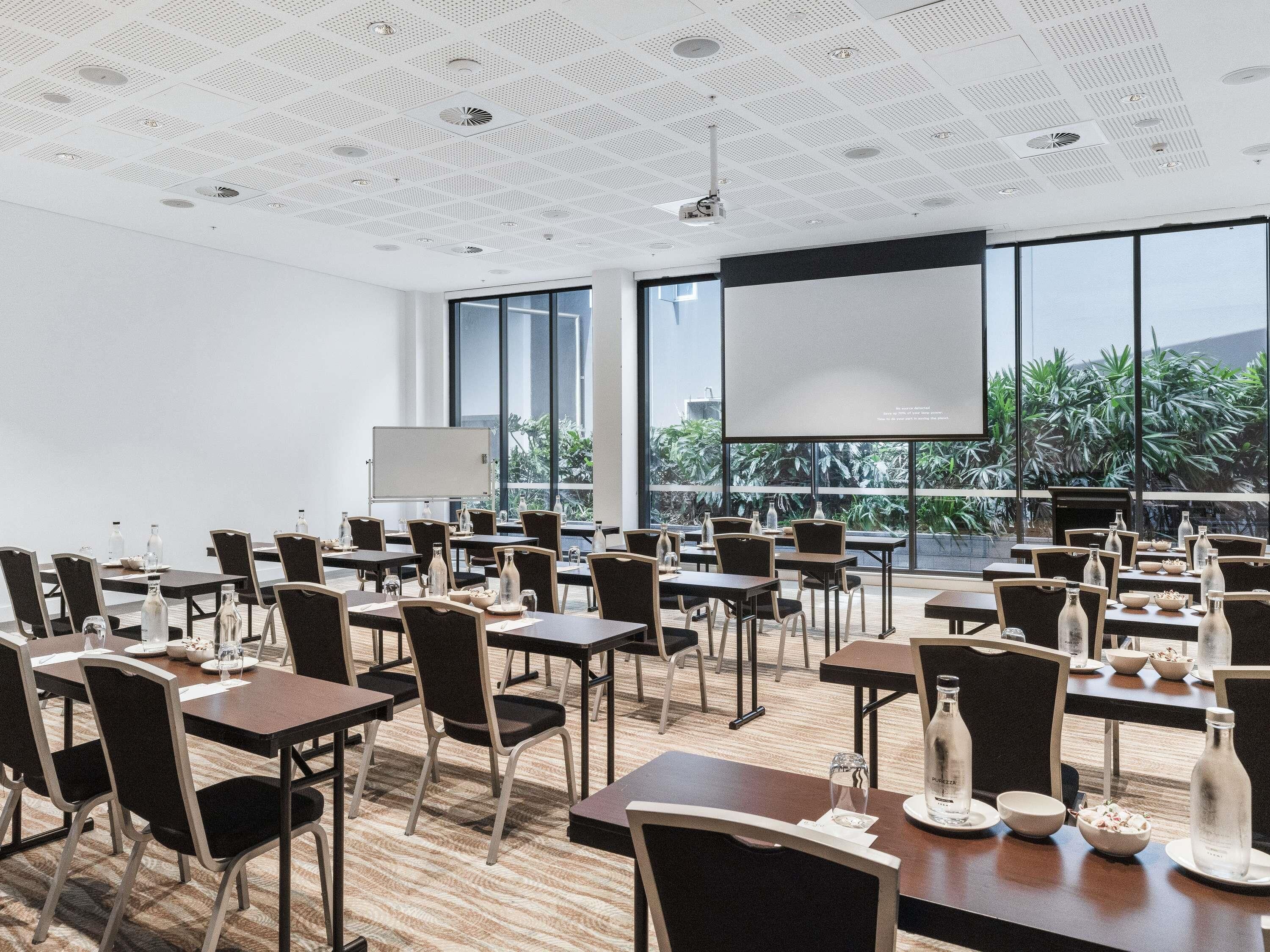 A conference room with tables, chairs, a projector screen, and large windows with a view of greenery.