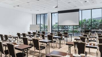 A conference room with tables, chairs, a projector screen, and large windows with a view of greenery.