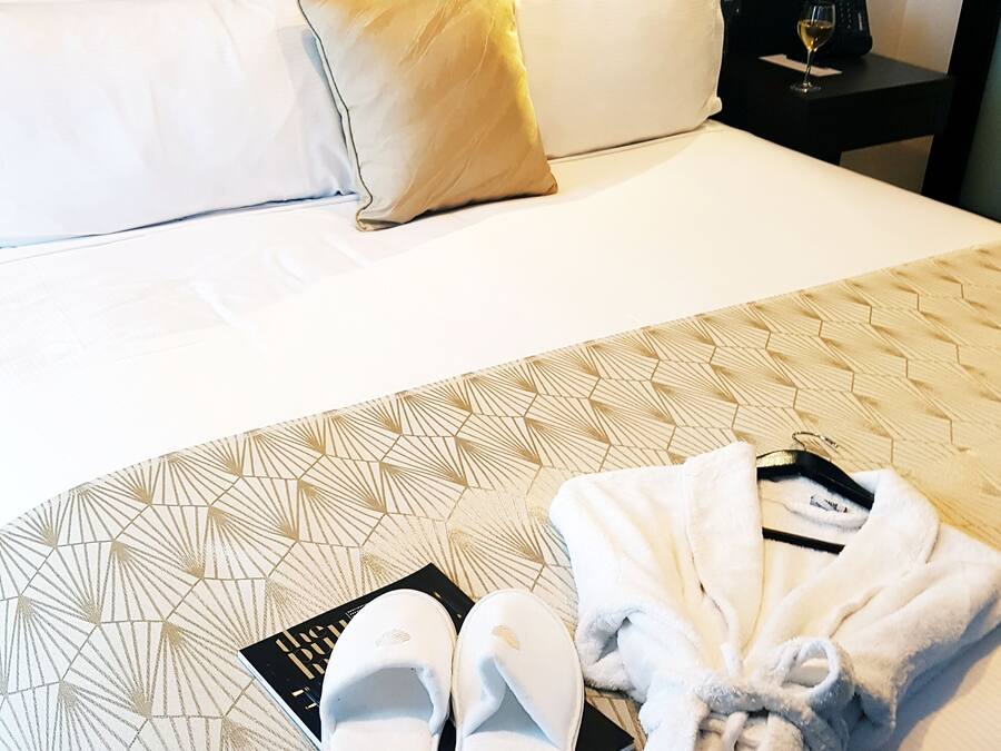 Cama bem arrumada com almofadas brancas e douradas, um par de chinelos brancos e um roupão sobre um edredom estampado.