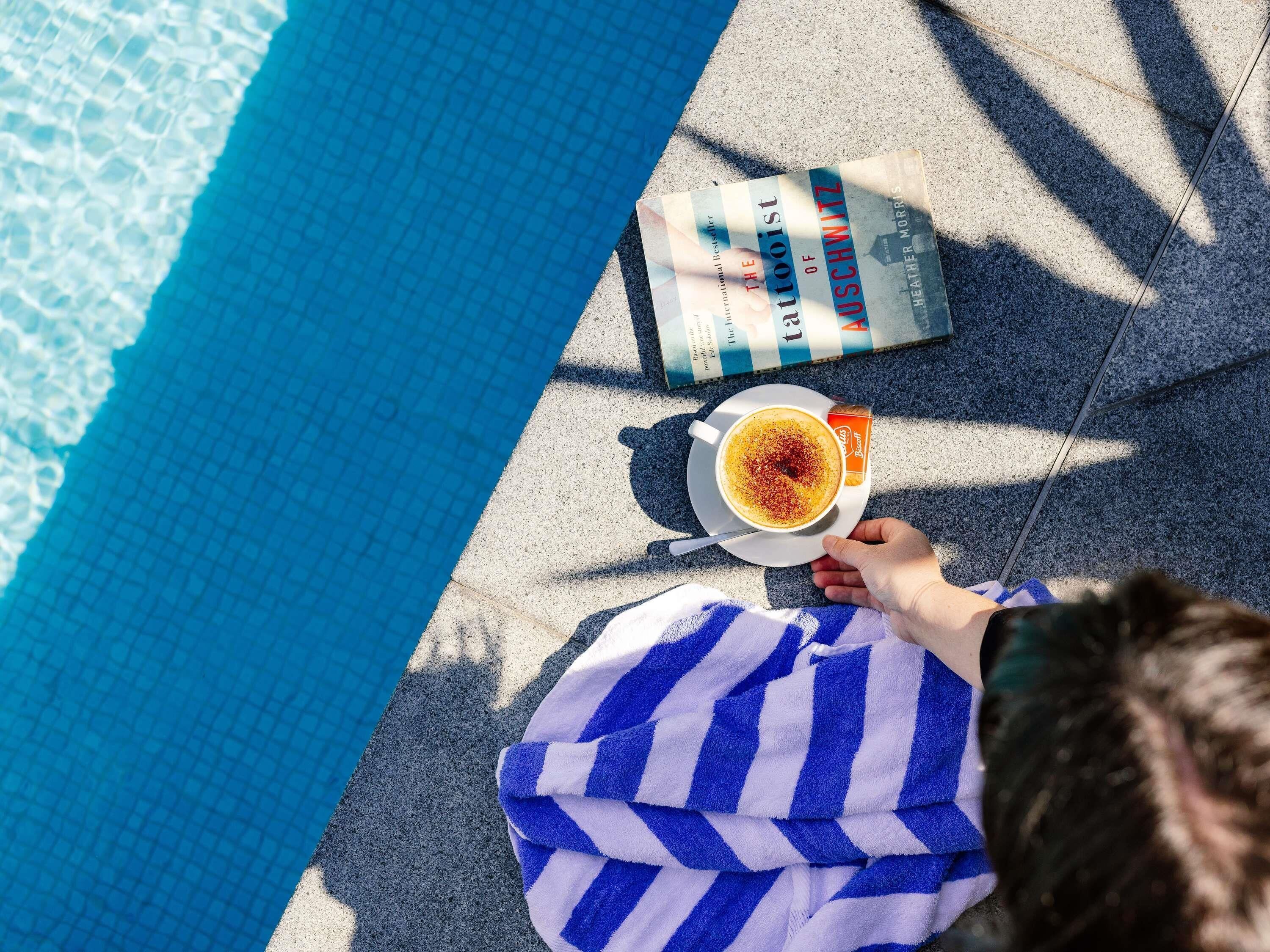 A person relaxing by a pool with a book and a cup of coffee.