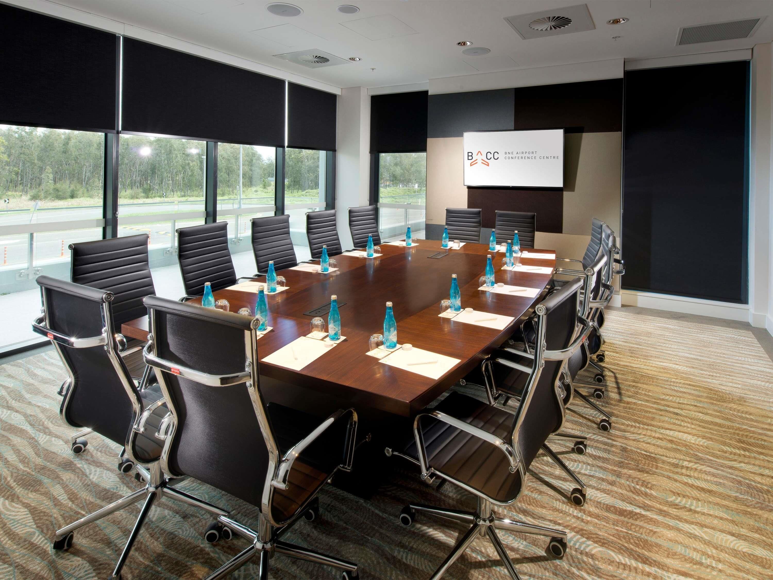 A conference room with a long table, chairs, and a projector screen displaying 'BACC BNE AIRPORT CONFERENCE CENTRE'.