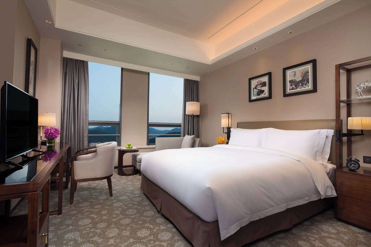 A hotel room with a large bed, a desk with a TV, a chair, a nightstand with a lamp, and a view of the mountains through the window.
