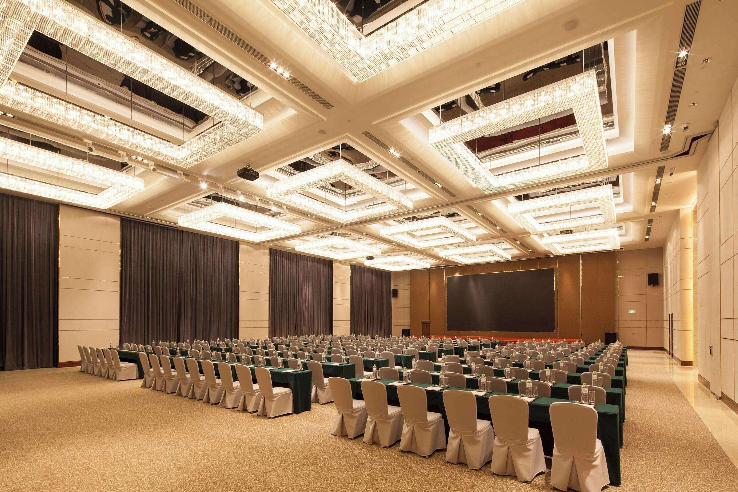 Une salle de conférence avec des rangées de tables et de chaises, un grand écran et un lustre au plafond.