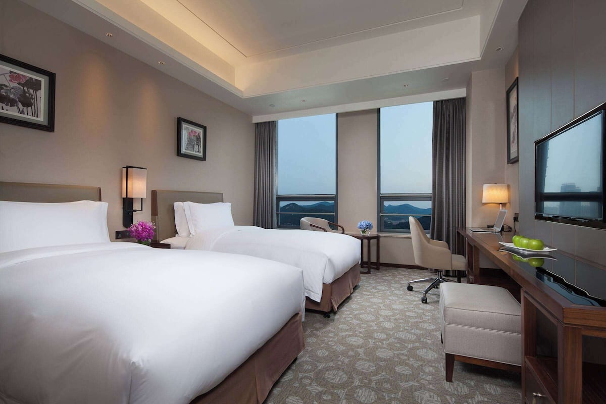 A hotel room with two beds, a desk, a chair, a TV, and a view of the mountains.