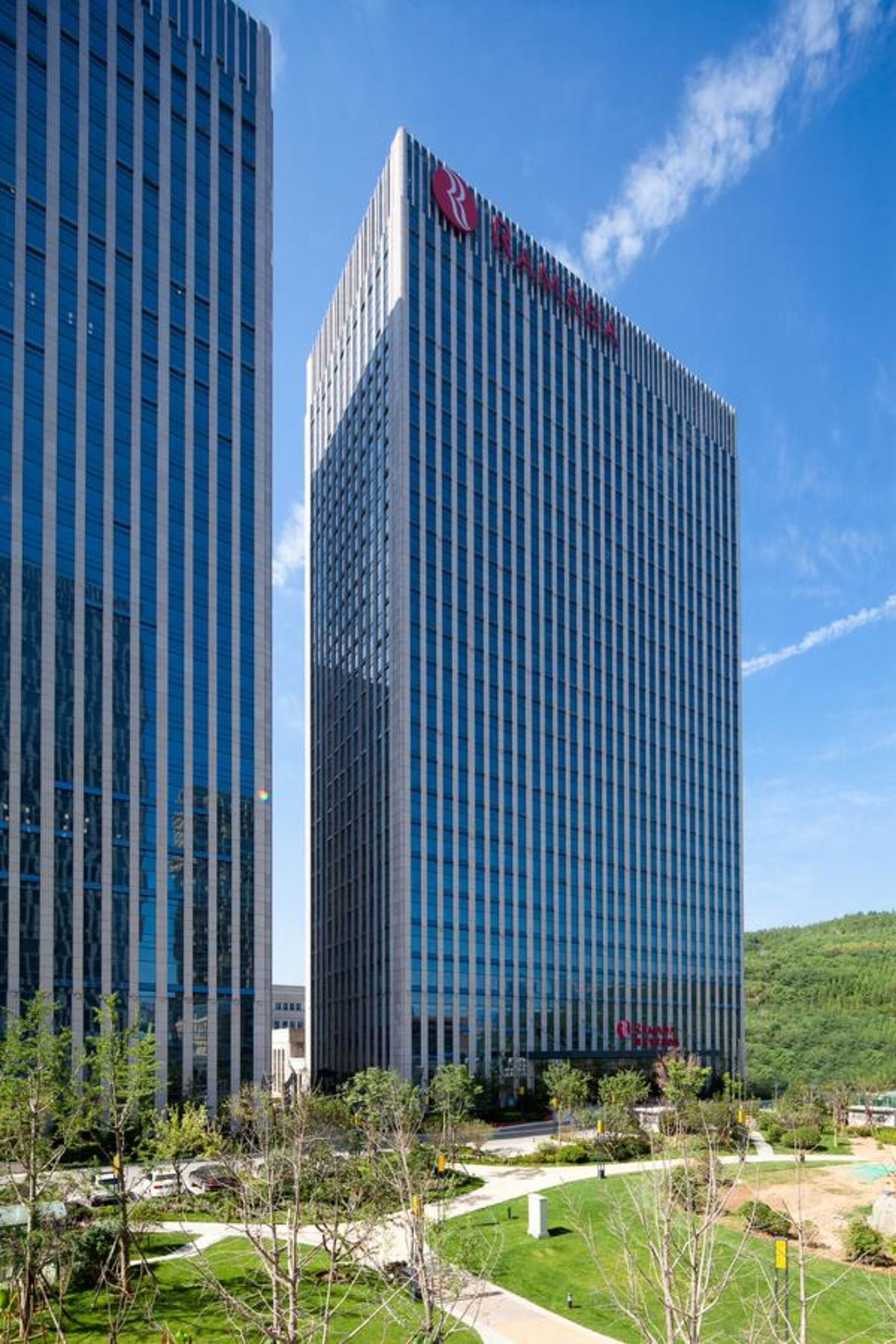 A modern building with a prominent 'Ramada' sign, surrounded by greenery and a clear sky.