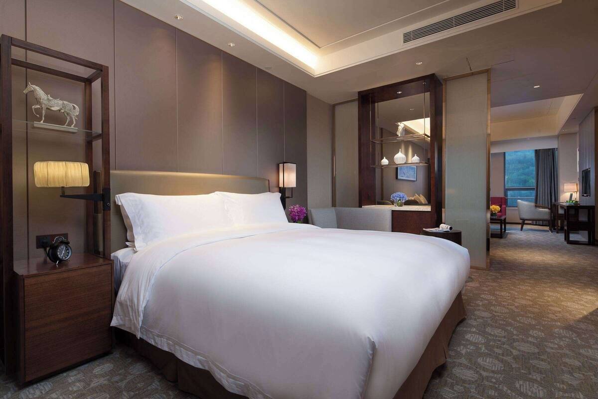 A hotel room with a large bed, a nightstand with a lamp, a mirror, and a desk with a chair.
