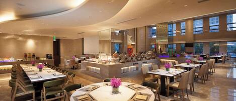 A modern restaurant with round tables and chairs, a central bar area, and large windows allowing natural light.