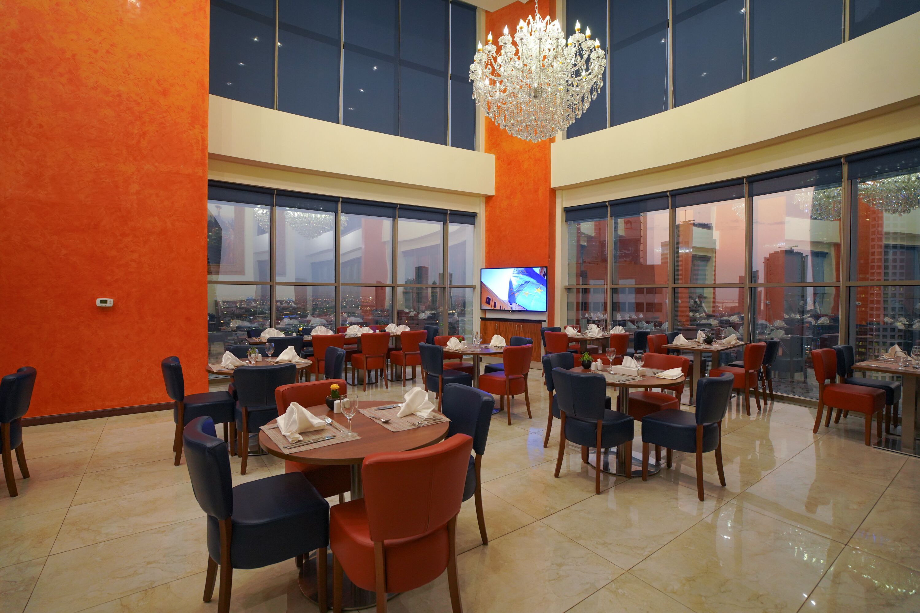 A spacious restaurant with high ceilings, large windows, and a modern chandelier. Tables are neatly arranged with chairs, and a television is mounted on the wall.