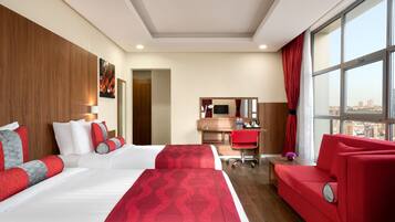 A hotel room with two beds, a desk, a red sofa, and a large window offering a city view.