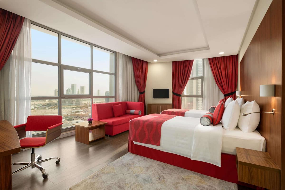 Habitación de hotel moderna con un ventanal grande, cortinas rojas, una cama con ropa blanca, un sofá rojo y un escritorio con una silla.