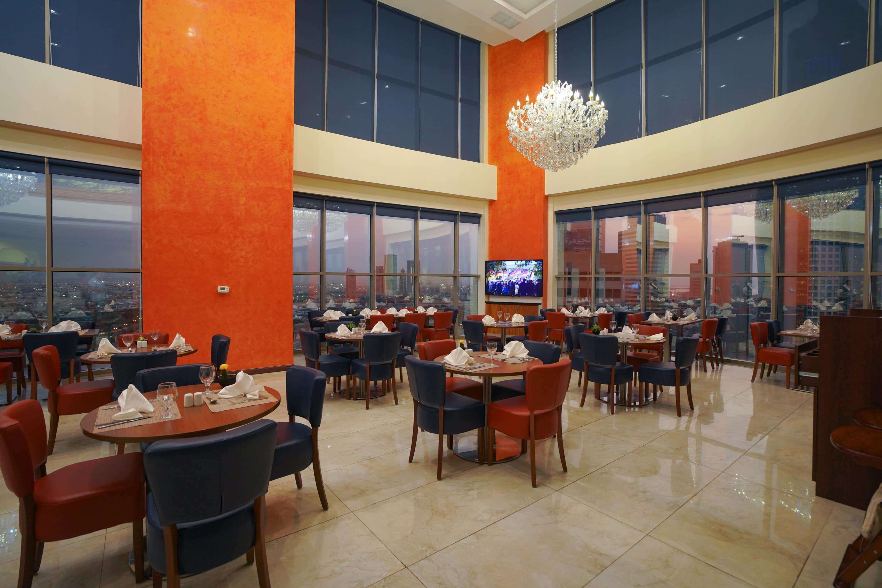 A spacious restaurant with high ceilings, large windows, and a city view. Tables are neatly arranged with chairs, and a chandelier hangs from the ceiling.
