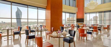 A modern dining area with round tables and chairs, large windows offering city views, and a chandelier.