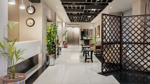 Een moderne hotellobby met een receptiebalie, potplanten en een zithoek met tafels en stoelen.