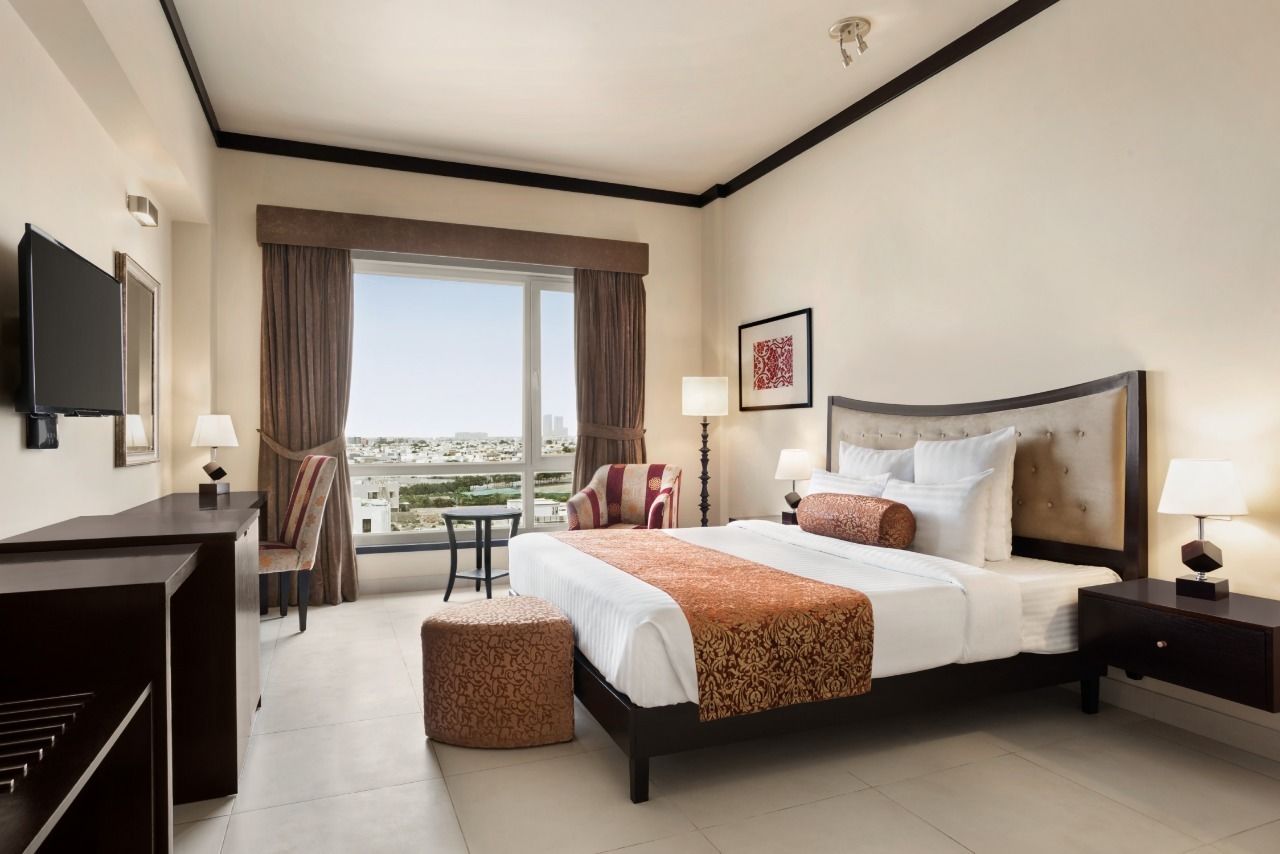 A hotel room with a large bed, a desk, a chair, a small table, and a view of the city through the window.