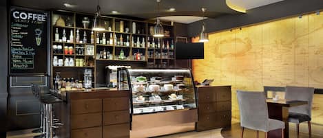 A coffee shop with a menu board, shelves of beverages, a display case with pastries, and a seating area with chairs and a table.