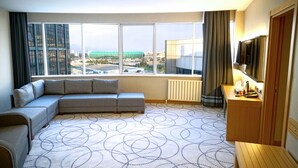 Ett modernt hotellrum med ett stort fönster som erbjuder utsikt över staden, en grå hörnsoffa, ett skrivbord i trä och en platt-TV.