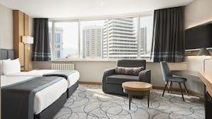 Ett hotellrum med två sängar, ett skrivbord, en stol, ett litet bord och ett stort fönster med gardiner.