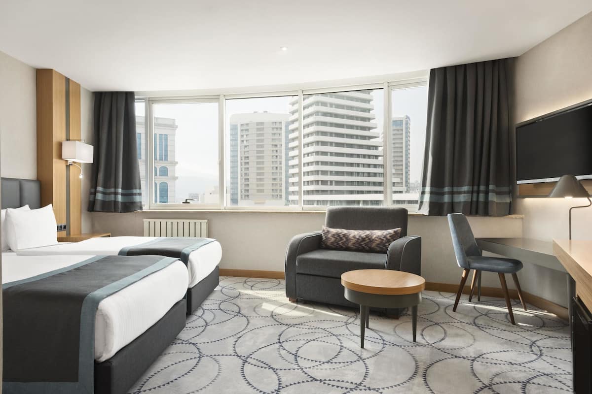 Ein Hotelzimmer mit zwei Betten, einem Schreibtisch, einem Stuhl, einem kleinen Tisch und einem großen Fenster mit Vorhängen.