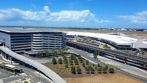 Una moderna terminal aeroportuaria con una estructura de estacionamiento de varios niveles, un puente peatonal y una zona ajardinada con árboles.