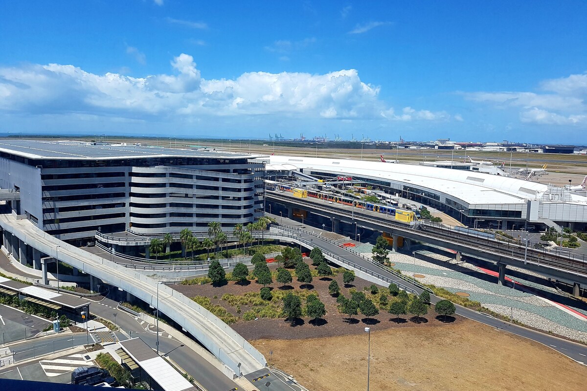 Una moderna terminal aeroportuaria con una estructura de estacionamiento de varios niveles, un puente peatonal y una zona ajardinada con árboles.