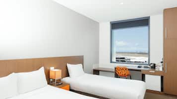 A hotel room with two beds, a desk, a chair, and a large window with a view of the airport.