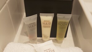 Un baño con una caja de pañuelos negros, tres artículos de tocador (cabello, cabello y cuerpo) y dos toallas blancas.