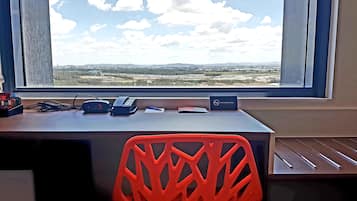 Un escritorio con vistas al paisaje que se ve por la ventana, una silla roja y algunos dispositivos electrónicos.